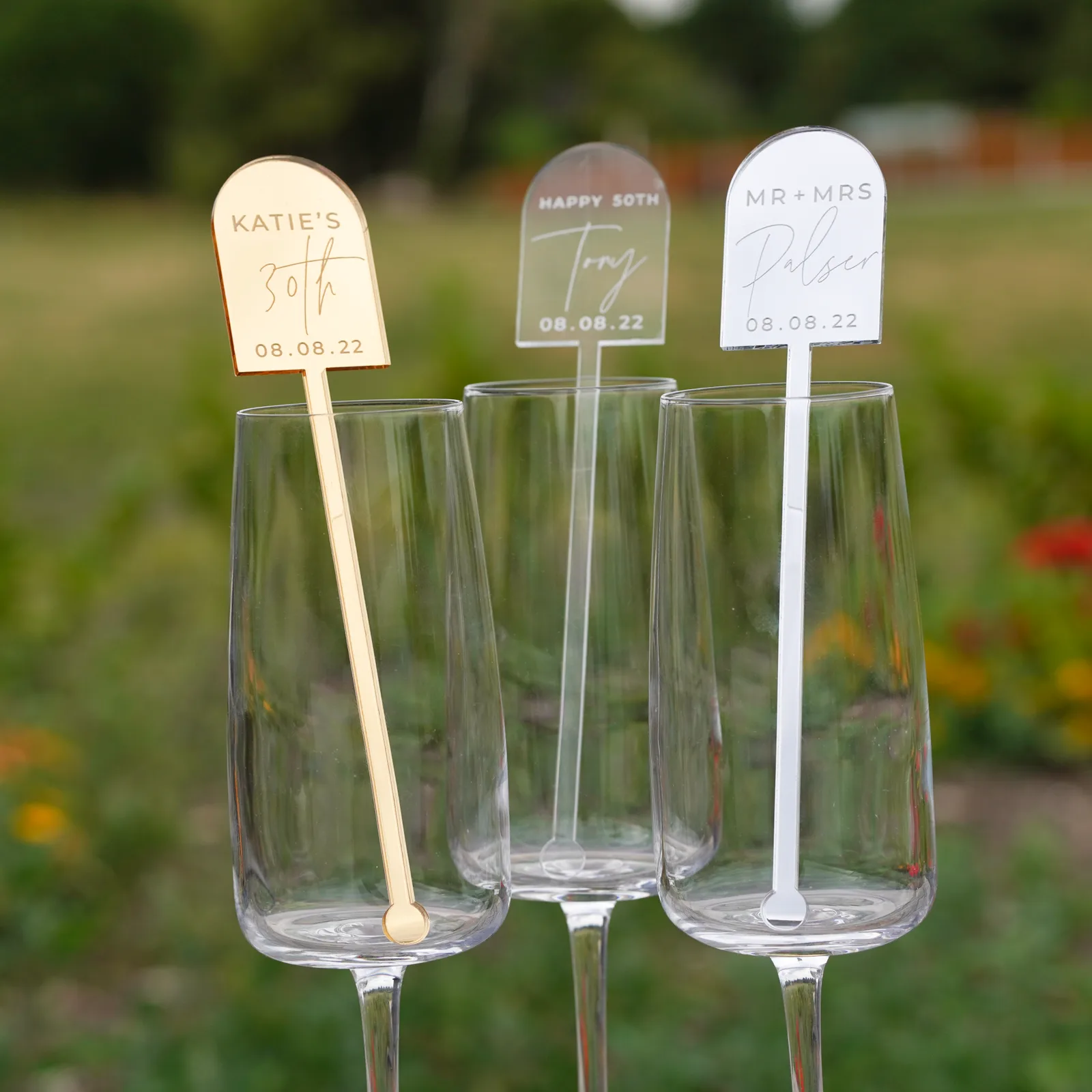 Three arched stirrers in mirror gold, clear and white acrylic, engraved with names and a date, standing in glasses